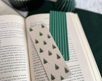 May include: A Christmas-themed bookmark with a repeating pattern of green Christmas trees on a cream background is placed in an open book. A second green bookmark with vertical lines is also visible. A dark green candle and a small Christmas tree are in the background.