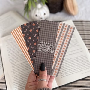 May include: A set of four rectangular bookmarks featuring various autumn-themed patterns including orange and white stripes, a dark brown background with small pumpkins, a dark grey gingham check with the text fall in love with reading, and pumpkins.