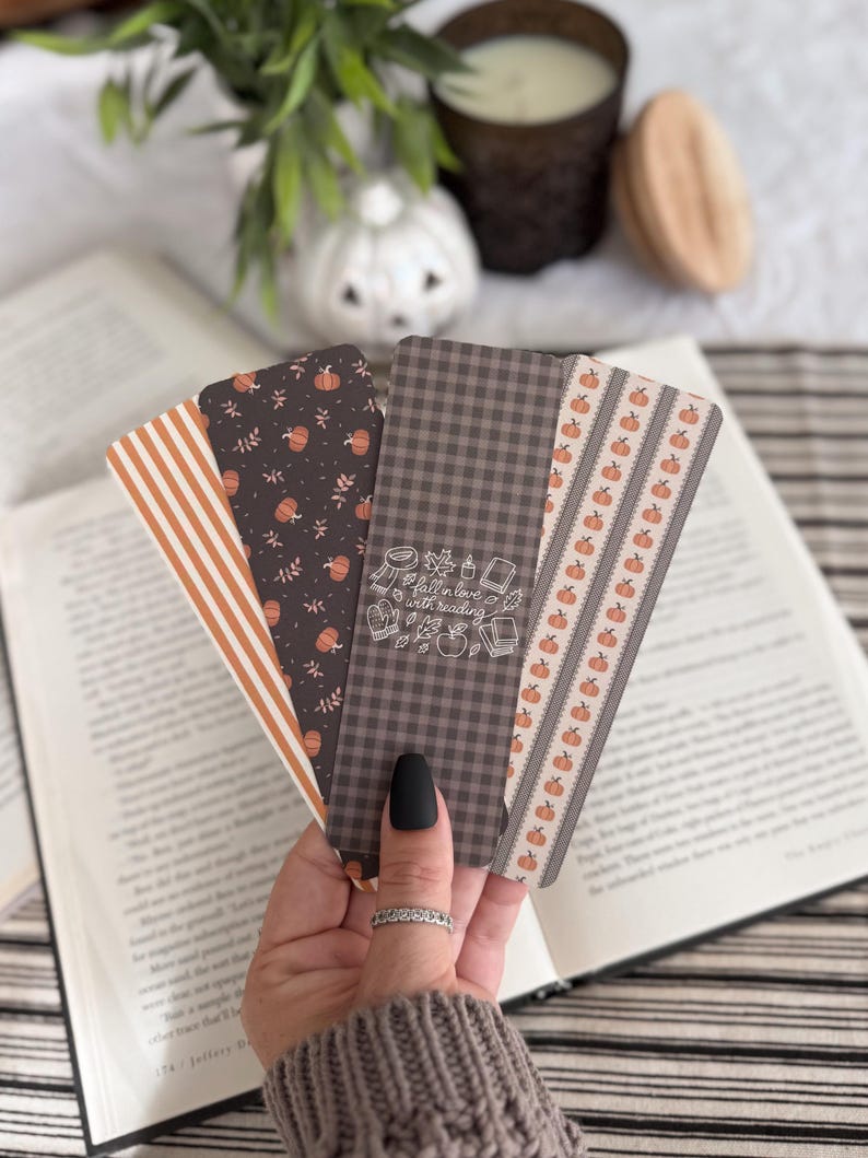 May include: A set of four rectangular bookmarks featuring various autumn-themed patterns including orange and white stripes, a dark brown background with small pumpkins, a dark grey gingham check with the text fall in love with reading, and pumpkins.