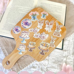 May include: A wooden cutting board with a variety of stickers on it. The stickers feature images of cakes, teacups, bunnies, and bouquets of flowers. The text "Look for the rest of the collection!" is printed on a pink background below the cutting board.