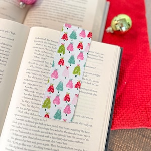 May include: A white bookmark with a repeating pattern of pink, green, and red Christmas trees. The trees are decorated with bows and have a white background.