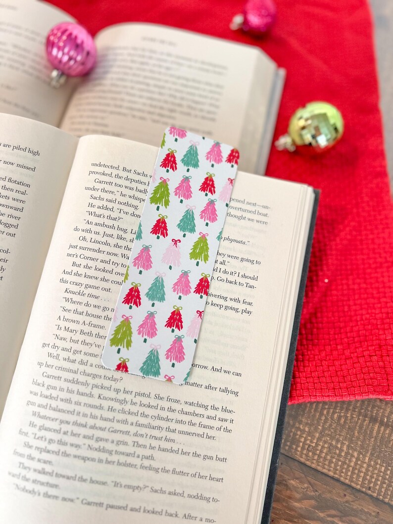 May include: A white bookmark with a repeating pattern of pink, green, and red Christmas trees. The trees are decorated with bows and have a white background.
