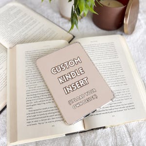 May include: A rectangular, light beige Kindle insert with rounded corners featuring the text CUSTOM KINDLE INSERT and (UPLOAD YOUR OWN DESIGN) in white, sans-serif lettering, resting on an open book with visible printed text.