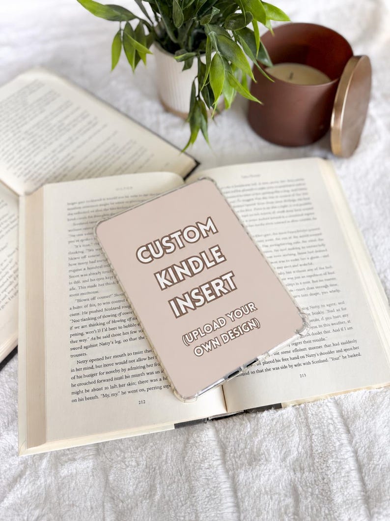 May include: A rectangular, light beige Kindle insert with rounded corners featuring the text CUSTOM KINDLE INSERT and (UPLOAD YOUR OWN DESIGN) in white, sans-serif lettering, resting on an open book with visible printed text.