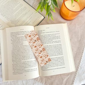 May include: An open book with a decorative bookmark featuring a pattern of books, coffee cups, and pastries. A lit orange candle and a small green plant are also visible, creating a cozy reading scene.