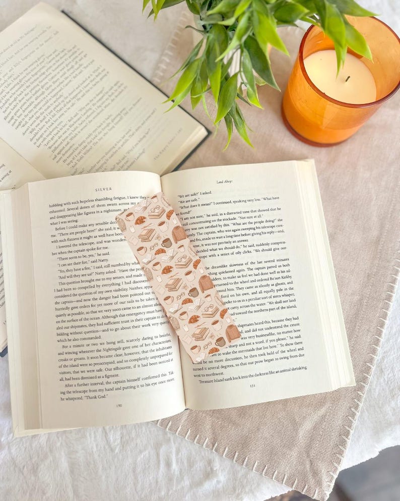May include: An open book with a decorative bookmark featuring a pattern of books, coffee cups, and pastries. A lit orange candle and a small green plant are also visible, creating a cozy reading scene.
