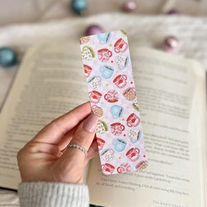 May include: A pink and white bookmark with a pattern of colorful mugs with snowflakes and other wintery designs. The bookmark is being held by a hand.