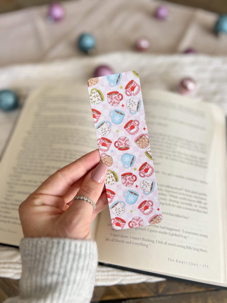 May include: A pink and white bookmark with a pattern of colorful mugs with snowflakes and other wintery designs. The bookmark is being held by a hand.