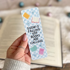 May include: A blue and white checkered bookmark with the text "SNOW IS FALLING and BOOKS ARE CALLING". The bookmark features illustrations of books with snowflakes.