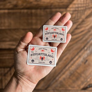 May include: Two white stickers with a red and black design. The stickers are shaped like a matchbox and say "SMUT BOARD" and "STFUATTDLAgg" with a skull and crossbones and a crescent moon.