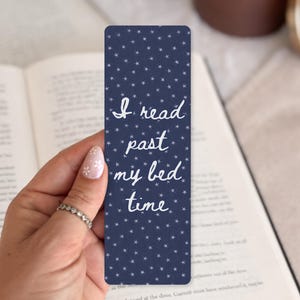 May include: A rectangular navy blue bookmark featuring a repeating pattern of small white stars. The text "I read past my bed time" is printed in a white, cursive script font in the center of the bookmark, oriented vertically.