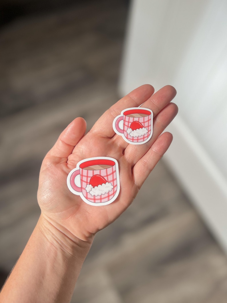 May include: Two pink and white mugs with a red Santa hat on top. The mugs are on a white background.