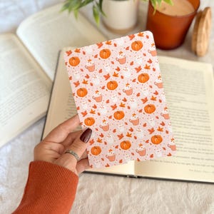 Pu&ograve; includere: Una carta rettangolare con un motivo ripetuto di zucche arancioni, fantasmi bianchi, tazze di bevande calde e foglie autunnali nei toni dell'arancione e del giallo su uno sfondo rosa chiaro, tenuta davanti a un libro aperto.