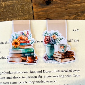 May include: Two watercolor bookmarks featuring a stack of books, a cup of coffee, and a vase of flowers. The bookmarks are on a page of a book with the text "We've won the" and "e're n" visible.