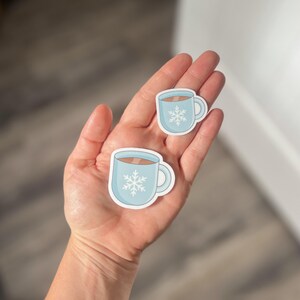 May include: Two blue and white coffee mugs with a snowflake design. The mugs are on a hand.