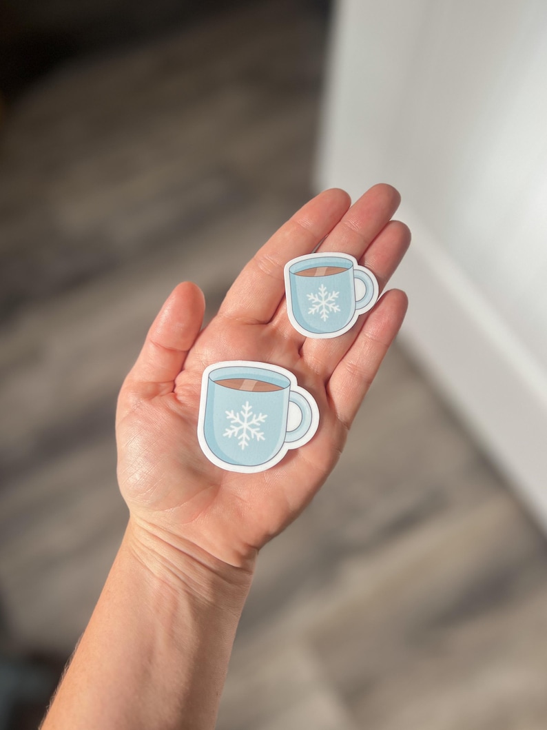 May include: Two blue and white coffee mugs with a snowflake design. The mugs are on a hand.