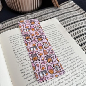 May include: A pink Halloween-themed bookmark with various framed illustrations, including pumpkins, ghosts, and candy. The word "SPOOK" is printed on the bookmark. The bookmark is placed in an open book.