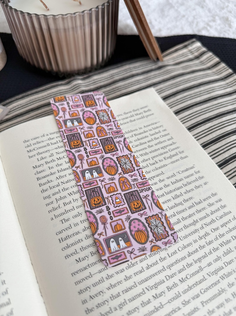 May include: A pink Halloween-themed bookmark with various framed illustrations, including pumpkins, ghosts, and candy. The word "SPOOK" is printed on the bookmark. The bookmark is placed in an open book.