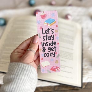 May include: Pink bookmark with a grid pattern and snowflakes. It features a mug of hot chocolate with marshmallows, a stack of books, and the text "Let's stay inside & get cozy".