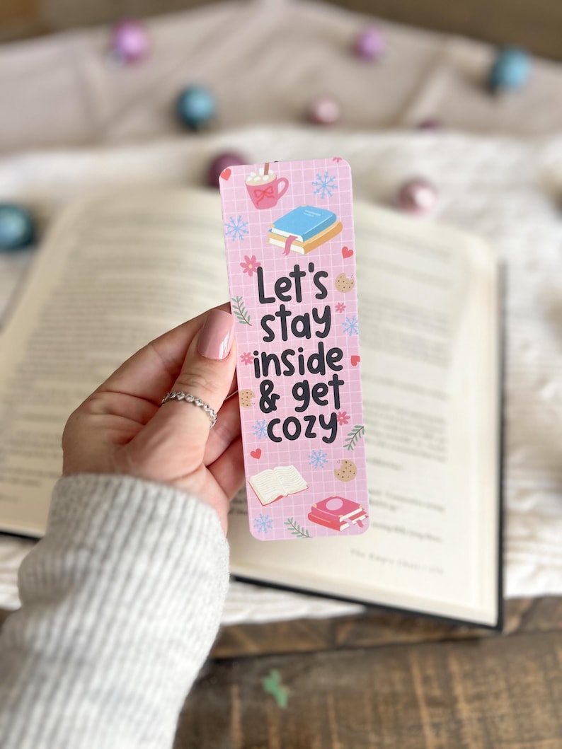 May include: Pink bookmark with a grid pattern and snowflakes. It features a mug of hot chocolate with marshmallows, a stack of books, and the text "Let's stay inside & get cozy".