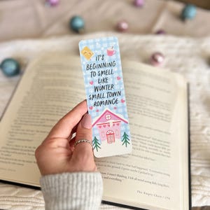 May include: A blue and white bookmark with a cute illustration of a house with a red door and snow-covered trees. The text on the bookmark reads "It's beginning to smell like winter small town romance."