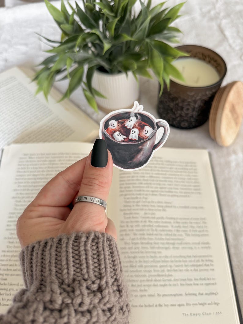 May include: A sticker featuring a black mug filled with a dark liquid and marshmallows with ghost faces. The sticker is held in front of an open book, with a plant and candle in the background.