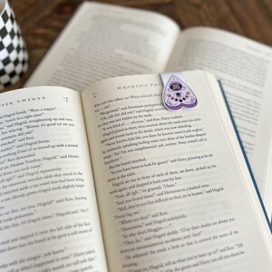 May include: An open book with a purple and black heart-shaped bookmark with a crystal design. The book is open to a chapter titled "Chapter Twenty" and the text reads "Hagrid's Tale".