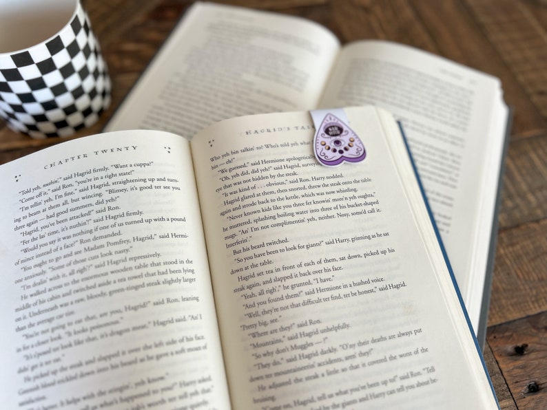 May include: An open book with a purple and black heart-shaped bookmark with a crystal design. The book is open to a chapter titled "Chapter Twenty" and the text reads "Hagrid's Tale".
