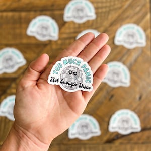 May include: A white sticker with a blue and white disco ball with a sad face and a tear. The text on the sticker reads "Too Much Panic Not Enough Disco".