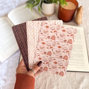 May include: Three rectangular cards with different designs are held in a hand. One card has a brown cable knit pattern, another has a cream cable knit pattern, and the third has a light pink background with a repeating pattern of books, sweaters, and coffee cups.