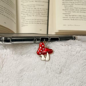 May include: A red and white mushroom charm with a gold-colored bail and clear rhinestones. The charm is attached to a clear phone case. The background includes open books and a white, fluffy surface.