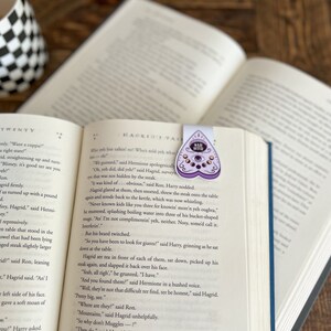 May include: A close-up of an open book with a purple heart-shaped bookmark featuring a Ouija board design. The bookmark is on the page with the text "Hagrid's Ta" visible. The book is open to a chapter titled "Chapter Twenty".