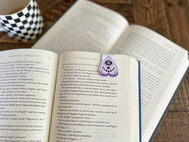 May include: A close-up of an open book with a purple heart-shaped bookmark featuring a Ouija board design. The bookmark is on the page with the text "Hagrid's Ta" visible. The book is open to a chapter titled "Chapter Twenty".