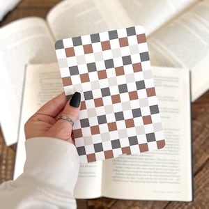 May include: A rectangular card featuring a repeating checkered pattern in alternating shades of dark gray, light gray, white, and muted terracotta brown. The card has rounded corners and is held against a background of open book pages.