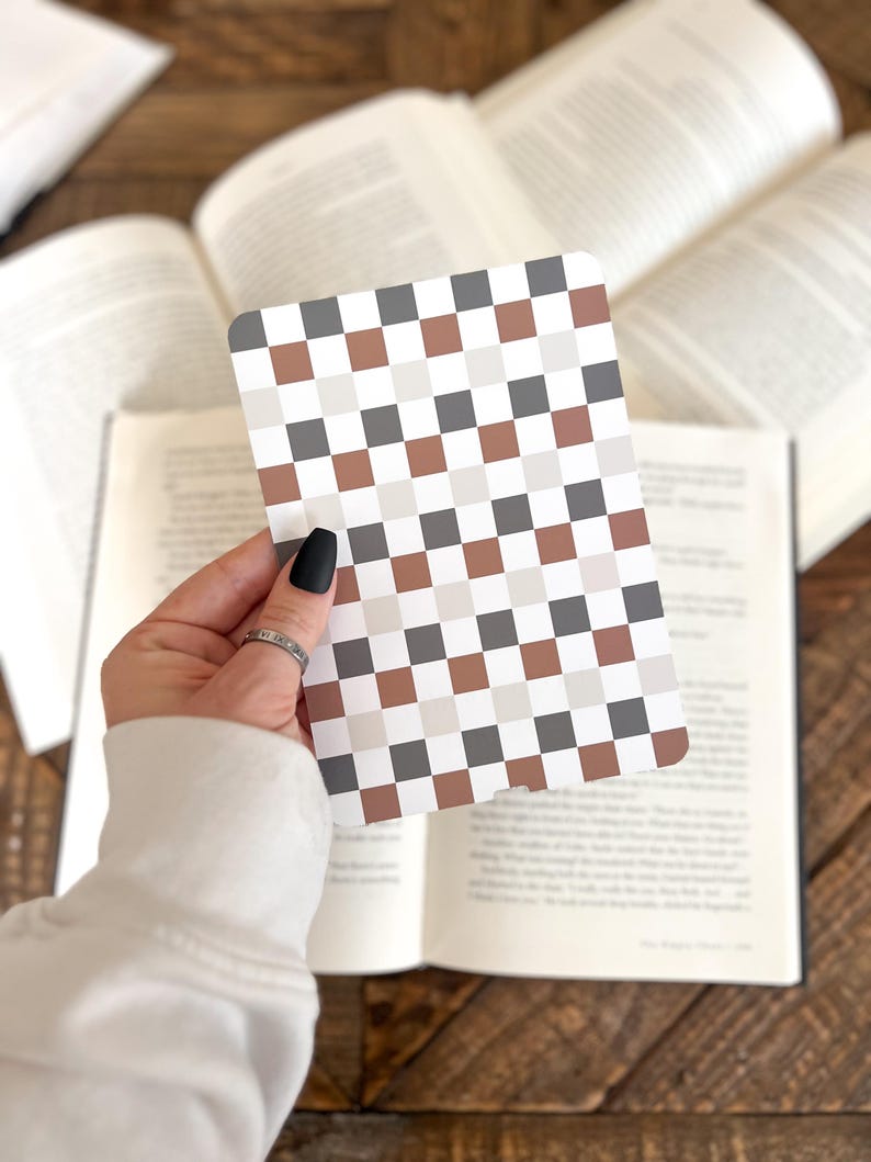 May include: A rectangular card featuring a repeating checkered pattern in alternating shades of dark gray, light gray, white, and muted terracotta brown. The card has rounded corners and is held against a background of open book pages.