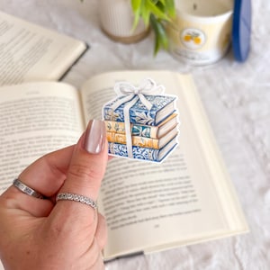May include: A sticker featuring a stack of books with blue and gold covers, tied with a white bow. The sticker is held in front of an open book, with a candle and plant in the background.