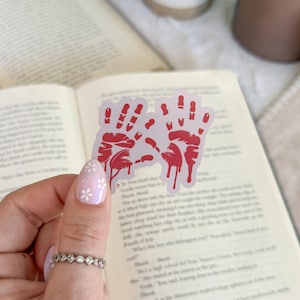 May include: A sticker featuring two red handprints with dripping blood detail. The sticker is held in front of an open book. The handprints are a spooky design.