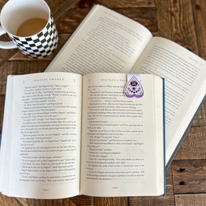 May include: An open book with a purple heart-shaped bookmark with a spooky eye design. The book is titled "Harry Potter and the Order of the Phoenix" and is open to Chapter Twenty. A mug with a black and white checkered design sits on a wooden table.