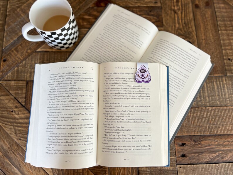 May include: An open book with a purple heart-shaped bookmark with a spooky eye design. The book is titled "Harry Potter and the Order of the Phoenix" and is open to Chapter Twenty. A mug with a black and white checkered design sits on a wooden table.