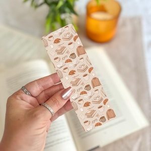 May include: A beige bookmark with a repeating pattern of books, coffee cups, eyeglasses, croissants, and sweaters. The bookmark is held over an open book. A candle and greenery are in the background.