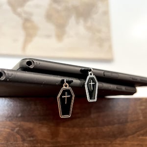May include: Two black coffin-shaped charms with a gold cross on each. The charms are hanging from a black tablet.