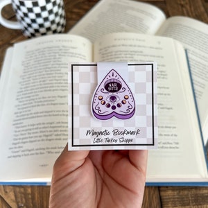 May include: Purple and white magnetic bookmark with a  'Nah, Bitch'  inscription inside a heart-shaped  ouija board design. The bookmark is on a white background with a black and white checkered pattern. The text 'Magnetic Bookmark' and 'Little Turkey Shoppe' is printed below the design.
