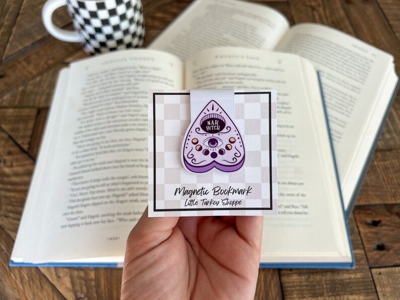May include: Purple and white magnetic bookmark with a  'Nah, Bitch'  inscription inside a heart-shaped  ouija board design. The bookmark is on a white background with a black and white checkered pattern. The text 'Magnetic Bookmark' and 'Little Turkey Shoppe' is printed below the design.