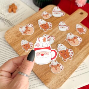 以下が含まれることがあります： 木製のまな板に貼られたクリスマスをテーマにしたステッカーのコレクション。ステッカーには、サンタクロースのマグカップ、ホイップクリームを添えたトナカイ、ジンジャーブレッドマンが描かれています。