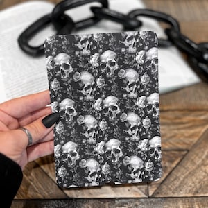 May include: A black and white notebook featuring a repeating pattern of skulls and roses. The notebook is held in front of a blurred background with a book and a black chain.