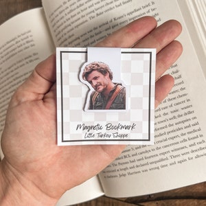 May include: A white magnetic bookmark with a black and white checkered background. The bookmark features a photo of a man with brown hair and a beard. The text "Magnetic Bookmark" and "Little Turkey Shoppe" is printed on the bookmark.