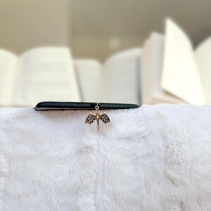 May include: A black smartphone sits on a white, fluffy surface, with a gold dragon charm hanging from it. The dragon has detailed wings and a small, yellow centre. Books are blurred in the background.