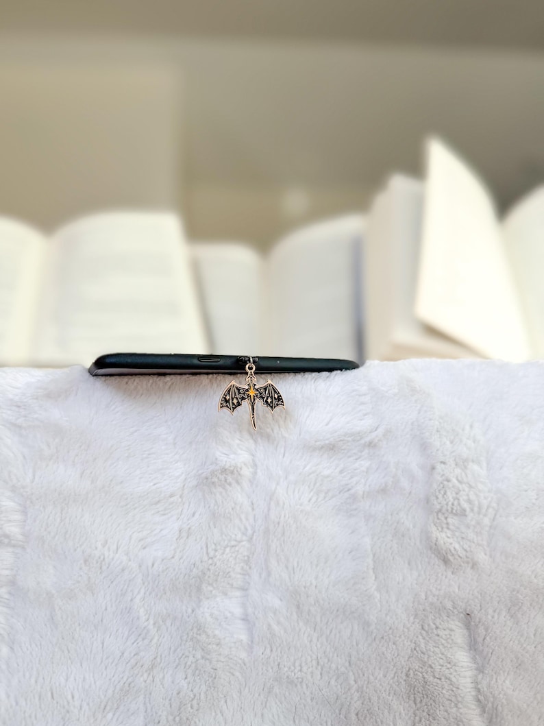 May include: A black smartphone sits on a white, fluffy surface, with a gold dragon charm hanging from it. The dragon has detailed wings and a small, yellow centre. Books are blurred in the background.