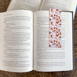May include: A floral bookmark with a white background and orange, pink, and brown flowers. The bookmark is made of paper and has a matte finish.