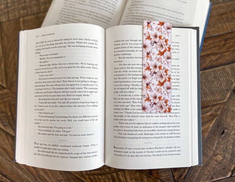 May include: A floral bookmark with a white background and orange, pink, and brown flowers. The bookmark is made of paper and has a matte finish.